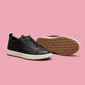 Black lace-up sneakers with white midsole and tan outsole on soft pink background